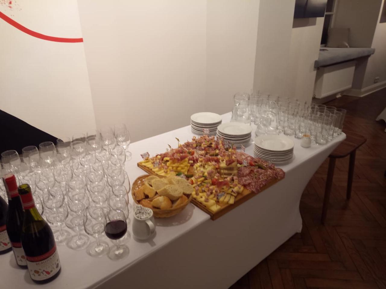 Stół z białym obrusem zastawiony przekąskami na drewnianej desce, pieczywem w koszyku, winem w butelkach i kieliszkach oraz stosem pustych talerzy i szklanek, przygotowany na przyjęcie.