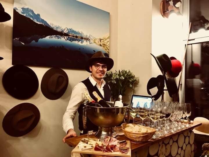 Uśmiechnięty mężczyzna w kapeluszu i kamizelce, za nim bar z przekąskami, winem i szampanem, w tle dekoracja z kapeluszy i obraz górskiego krajobrazu.