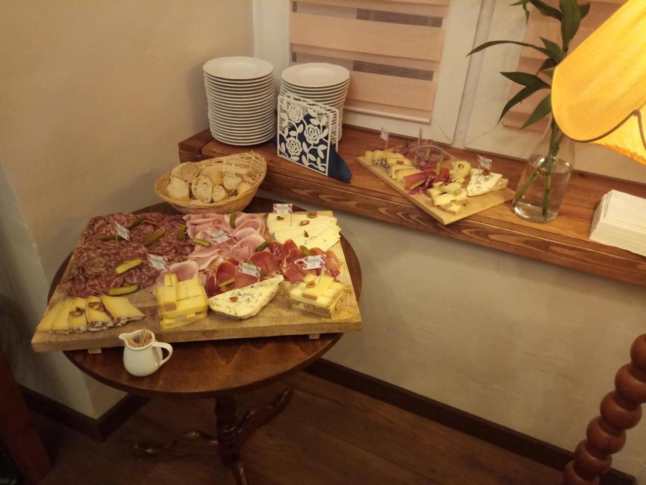 Deska serów i wędlin z dodatkami na drewnianym stole, obok stos talerzy i koszyk z pieczywem, całość w ciepłym oświetleniu.