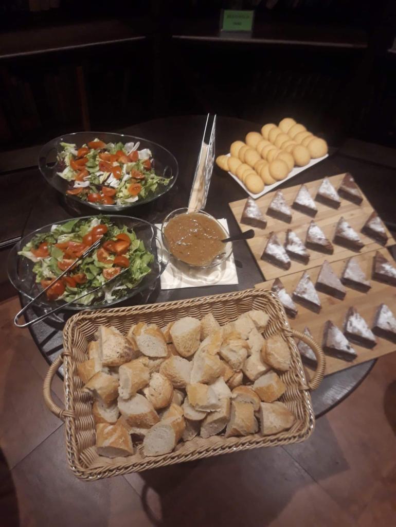 Bufet z sałatkami, pieczywem w koszyku, sosem, ciastkami i kawałkami ciasta posypanymi cukrem pudrem, ustawiony na ciemnym stole.