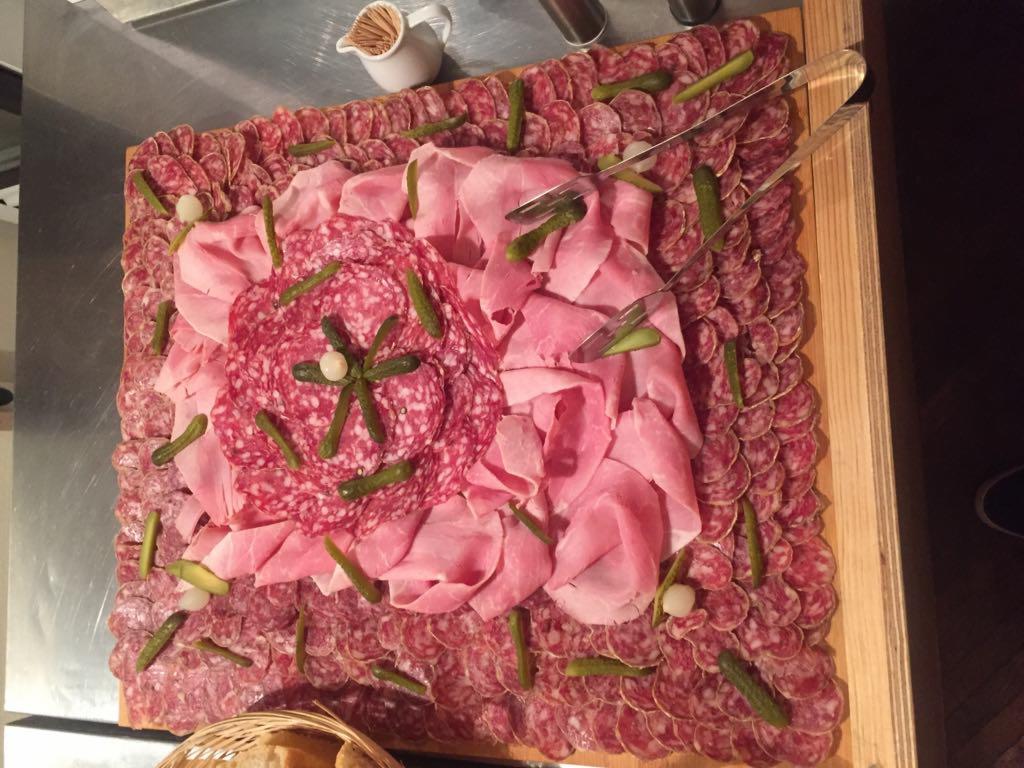 Dekoracyjna deska wędlin i pikli, ułożona na drewnianej tacy z metalowymi szczypcami, widoczny dzbanuszek na sos.