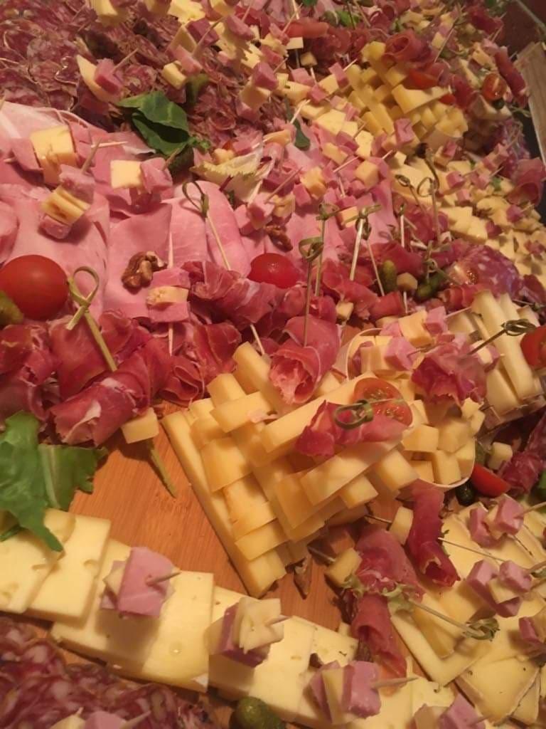 Bogato zastawiony stół z przekąskami na przyjęcie: szynka parmeńska, salami, sery żółte i pleśniowe, pomidorki koktajlowe, korniszony, orzechy włoskie, wszystko nabite na wykałaczki.