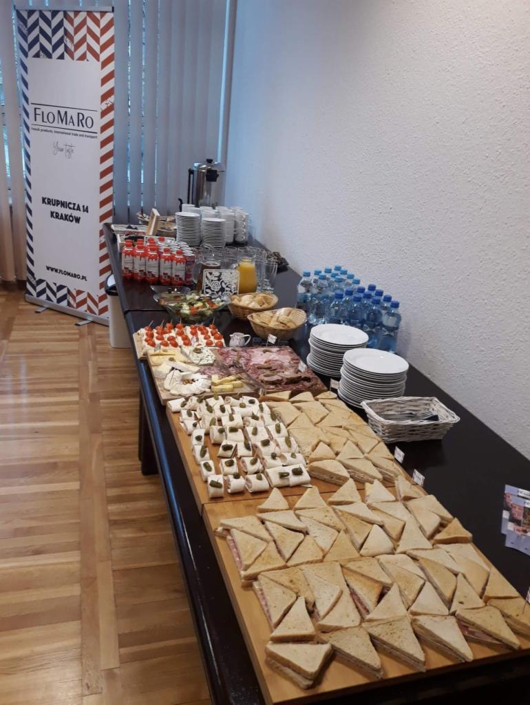 Stół z różnorodnym cateringiem: kanapki trójkątne, roladki z ogórkiem, deski serów i wędlin, napoje, w tle banner reklamowy Flomaro i biała ściana.