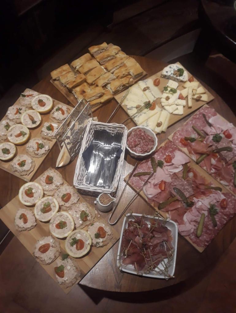 Stół z drewnianym blatem, na którym znajdują się drewniane deski z różnorodnymi przekąskami: kanapki, sery, wędliny, pasta na pieczywie z pomidorkiem koktajlowym, miseczka z salami, koszyk ze...