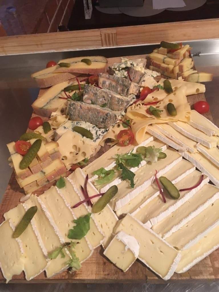 Deska serów z różnymi gatunkami, udekorowana pomidorkami koktajlowymi, ogórkami konserwowymi i zieleniną, ułożona na drewnianej desce.