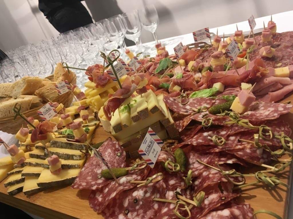 Bogato zastawiony stół z deską serów i wędlin, dekoracyjnie ułożonych na drewnianej desce, z flagami 'FROMARO' i kieliszkami w tle.