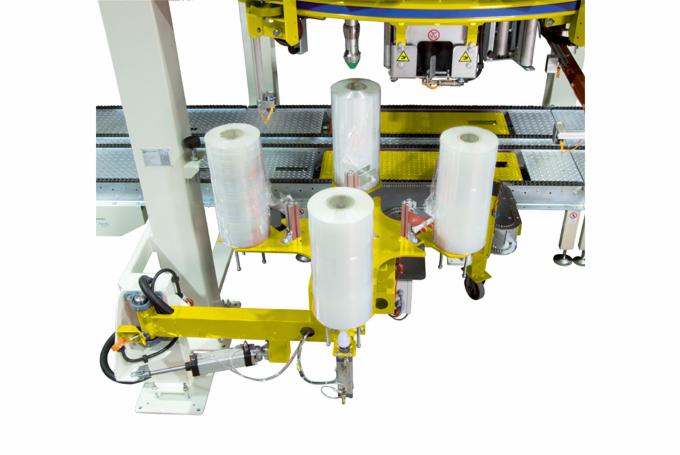 Automatyczna maszyna pakująca z rolkami folii termokurczliwej, żółte ramię robota z pneumatycznym siłownikiem, biała kolumna nośna, widok z góry na podajnik taśmowy.
