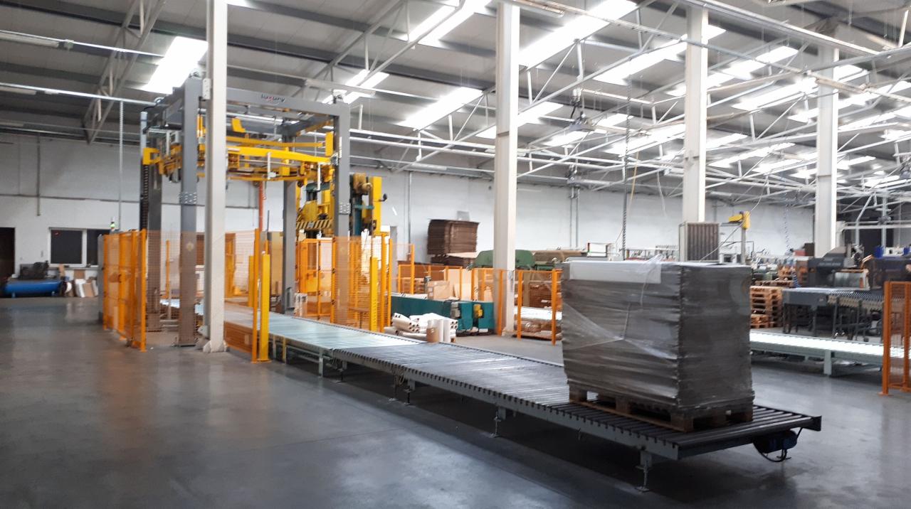 Automatyczna linia pakująca z owiniętą folią paletą na transporterze w hali produkcyjnej, z widocznym żółtym robotem pakującym i zabezpieczeniami.