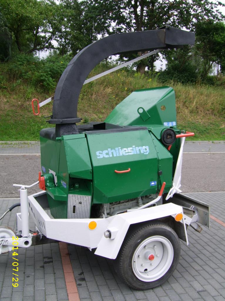Zielona rębarka do gałęzi marki Schliesing na przyczepie z białym podwoziem, zaparkowana na kostce brukowej. Widoczne drzewa w tle, urządzenie po deszczu.