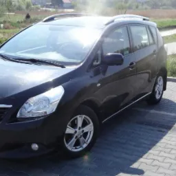 Czarny minivan Toyota Verso na parkingu z kostki brukowej, w tle rzeka, most i zieleń.