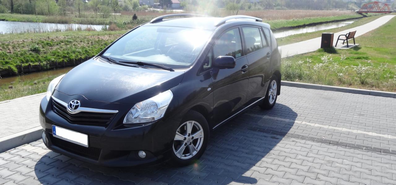 Czarny minivan Toyota Verso na parkingu z kostki brukowej, w tle rzeka, most i zieleń.