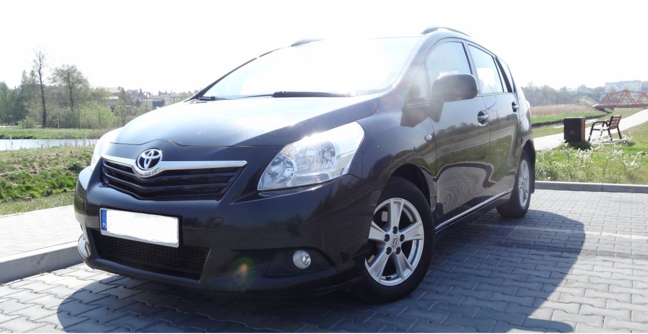 Czarny samochód osobowy Toyota Verso na brukowanym parkingu w słoneczny dzień, z widocznym fragmentem krajobrazu z jeziorkiem i czerwonym mostkiem w tle.