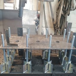 Usługi ślusarsko-spawalnicze Rafał Hałupka - Metalowa konstrukcja z gwintowanymi prętami i przyspawanymi elementami, ustawiona na perforowanym stole warsztatowym. W tle narzędzia i akcesoria.