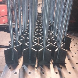 Usługi ślusarsko-spawalnicze Rafał Hałupka - Metalowe elementy konstrukcyjne z gwintowanymi prętami, spawane na perforowanym stole spawalniczym. Widoczne spawarki IDEAL w tle. Precyzyjne wykonanie.