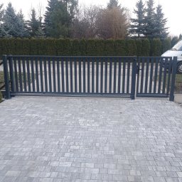 Usługi ślusarsko-spawalnicze Rafał Hałupka - Nowoczesna, przesuwna brama wjazdowa z pionowymi szczeblami, szara kolorystyka, widoczny mechanizm otwierania, kostka brukowa na podjeździe, tło z zielonych krzewów i drzew.