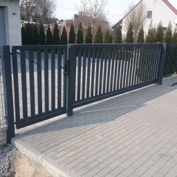 Usługi ślusarsko-spawalnicze Rafał Hałupka - Nowoczesna, przesuwna brama wjazdowa w kolorze grafitowym, z pionowymi szczeblami, zamontowana na posesji z kostką brukową i zielonymi tujami w tle.