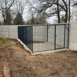 Usługi ślusarsko-spawalnicze Rafał Hałupka - Nowoczesna, metalowa brama wjazdowa z pionowymi prętami, osadzona w betonowym fundamencie, na tle betonowego ogrodzenia panelowego. Surowy krajobraz, pochmurne niebo.