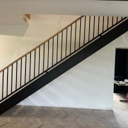 Usługi ślusarsko-spawalnicze Rafał Hałupka - Nowoczesne schody z czarną metalową konstrukcją i drewnianą poręczą, widoczne na tle białej ściany i podłogi z płytek w jodełkę. Minimalistyczny design.