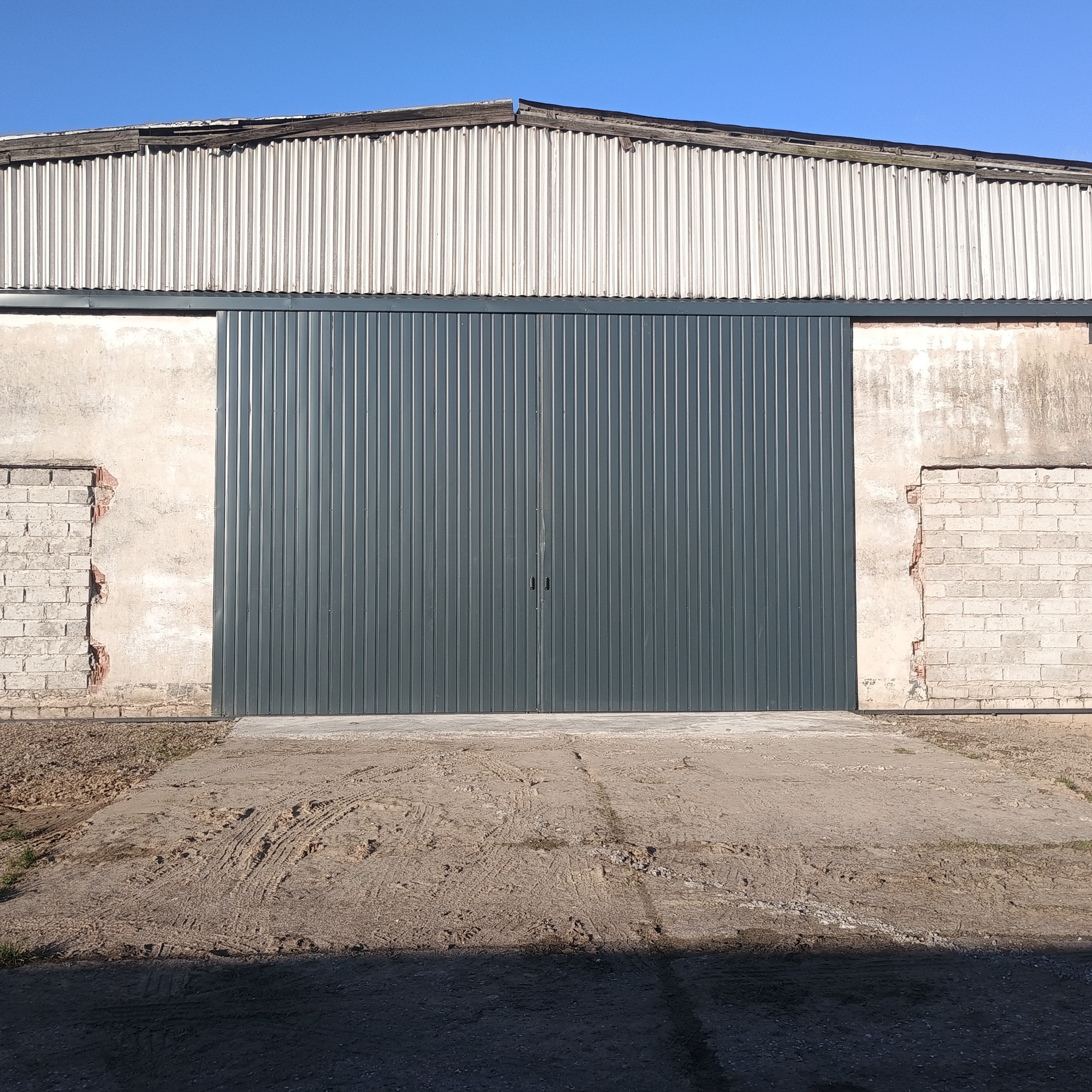 Szara, dwuskrzydłowa brama wjazdowa z blachy trapezowej, zamontowana w starej, częściowo zniszczonej hali z elementami muru ceglanego po bokach.