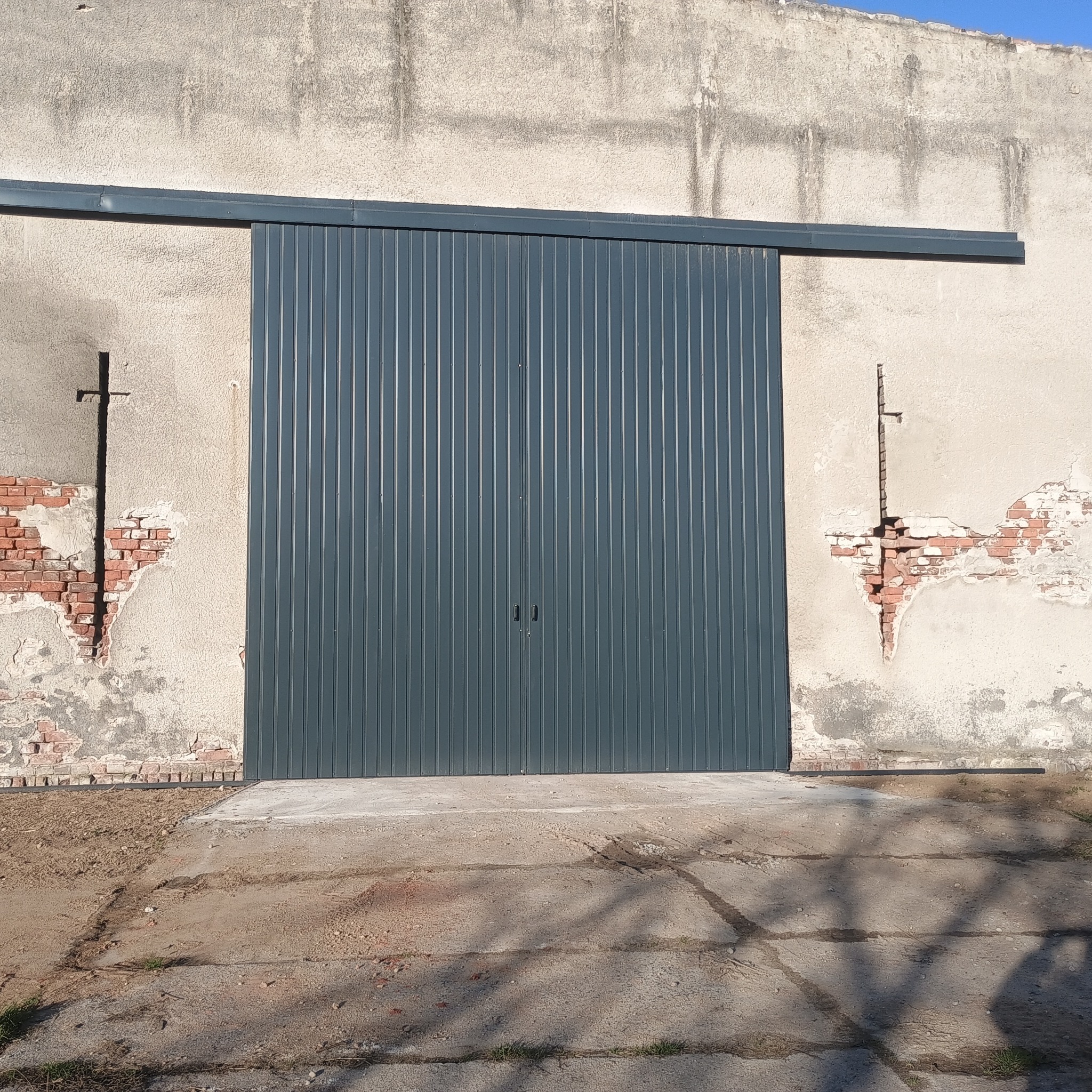 Szara, przesuwna brama z pionowymi panelami, zamontowana na tle starego, otynkowanego budynku z fragmentami odsłoniętej cegły. Widoczne ślady po dawnych elementach.