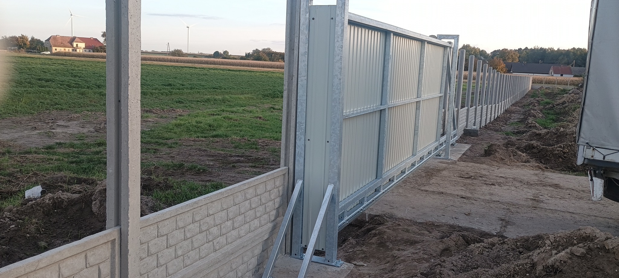 Nowoczesna brama przesuwna z metalowymi panelami, widoczna z boku, z betonową podmurówką i słupkami. W tle krajobraz wiejski z turbiną wiatrową.