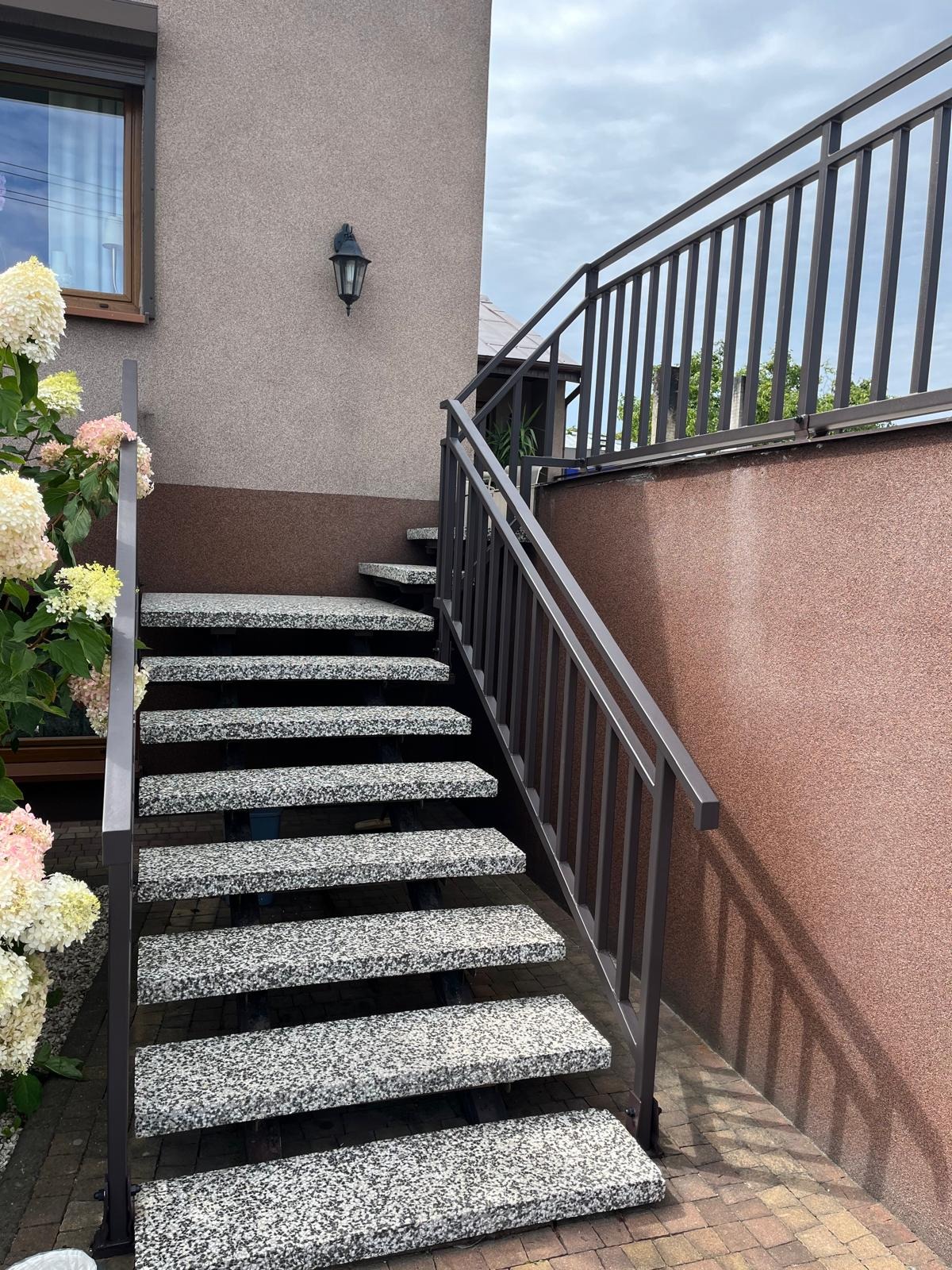 Zewnętrzne schody z szarego lastryko z metalową balustradą w kolorze grafitowym, prowadzące do podwyższonego tarasu. Obok rosną hortensje.