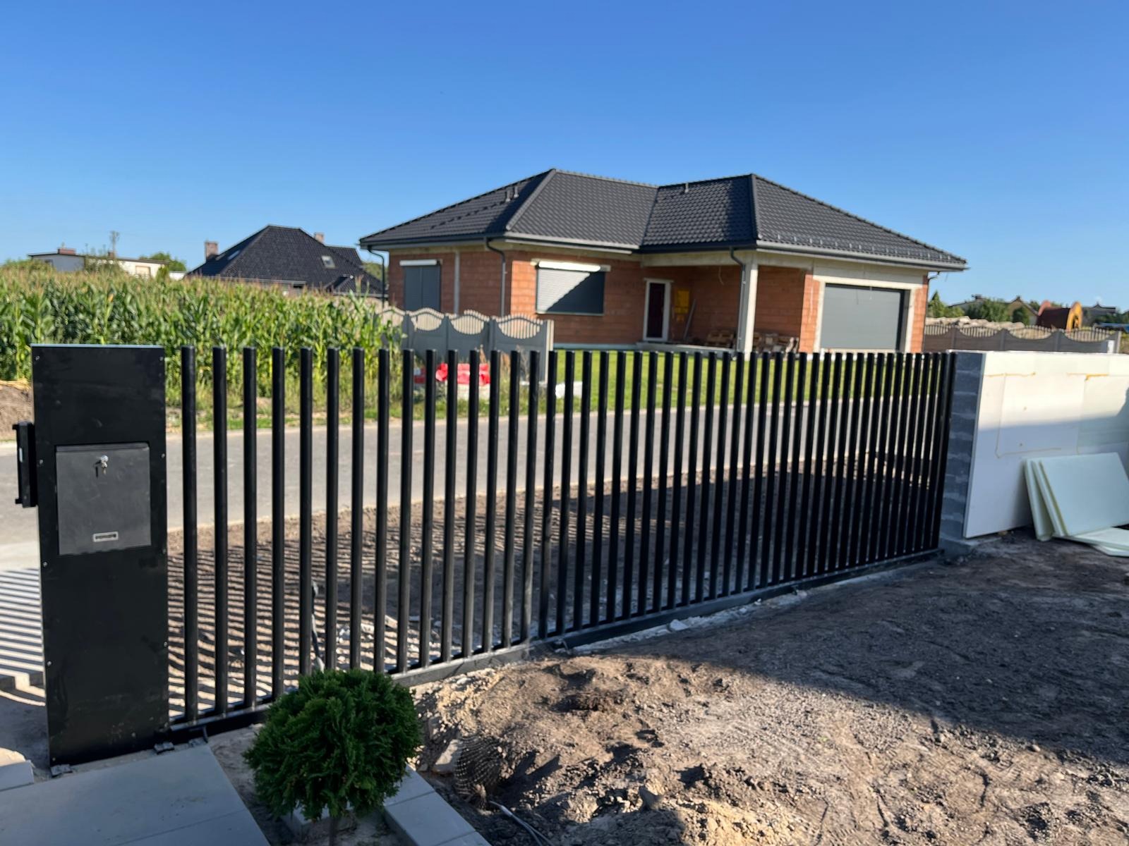 Nowoczesne, czarne ogrodzenie metalowe z pionowych prętów, z furtką i skrzynką na listy, na tle domu jednorodzinnego w słoneczny dzień. Minimalistyczny design.