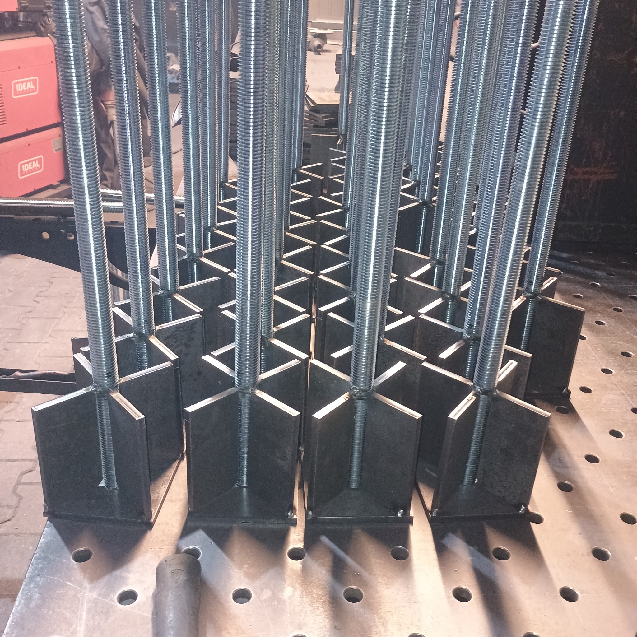 Metalowe elementy konstrukcyjne z gwintowanymi prętami, spawane na perforowanym stole spawalniczym. Widoczne spawarki IDEAL w tle. Precyzyjne wykonanie.