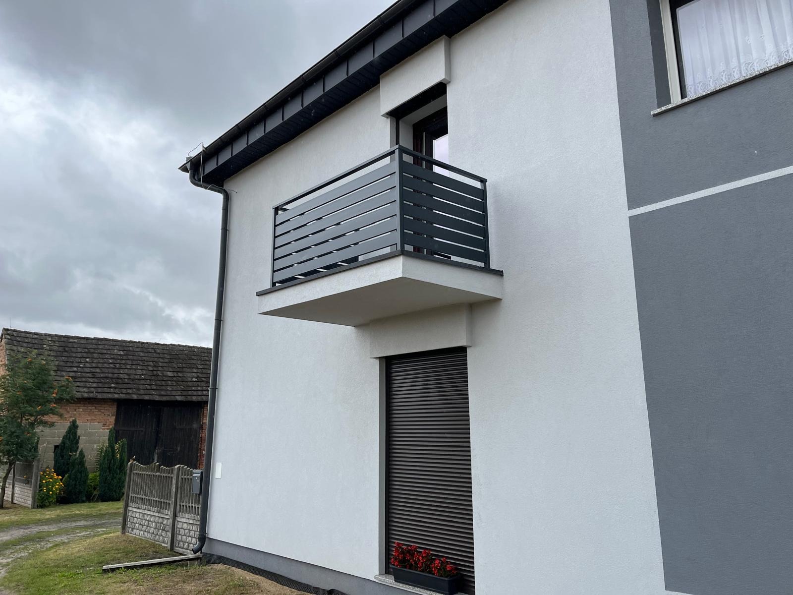 Nowoczesna, pozioma, metalowa balustrada balkonowa w kolorze antracytu, zamontowana na białym budynku z szarą wstawką. W tle widoczny budynek gospodarczy.