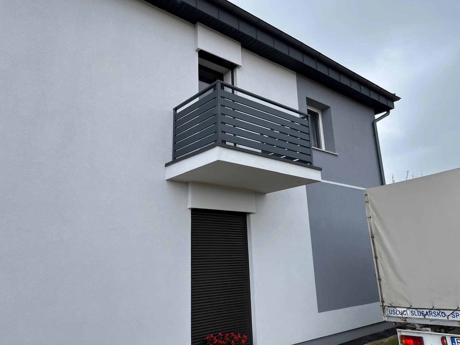 Nowoczesna, pozioma balustrada balkonowa w kolorze antracytowym, zamontowana na balkonie budynku o dwukolorowej elewacji. Widok z dołu, fragment ciężarówki w tle.