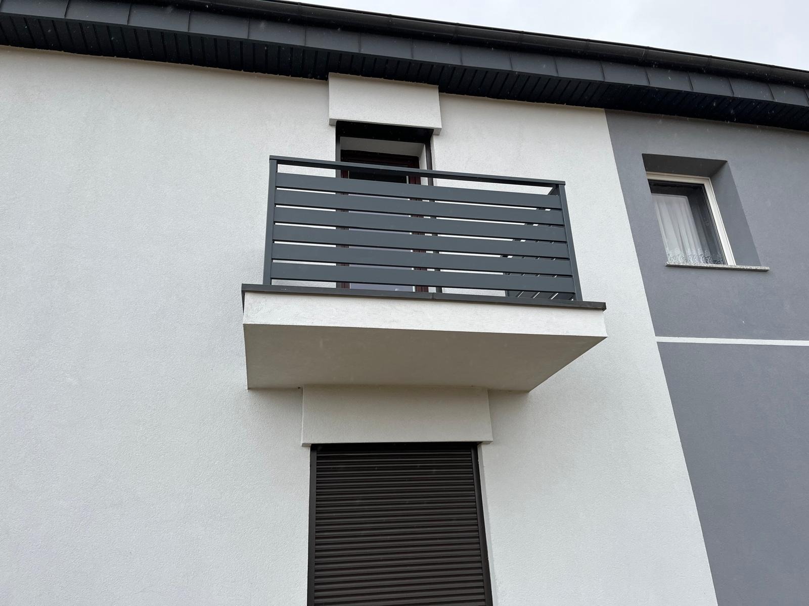 Nowoczesna, pozioma balustrada balkonowa w kolorze antracytowym, zamontowana na białym budynku z szarą elewacją. Minimalistyczny design, proste linie, widok z dołu.