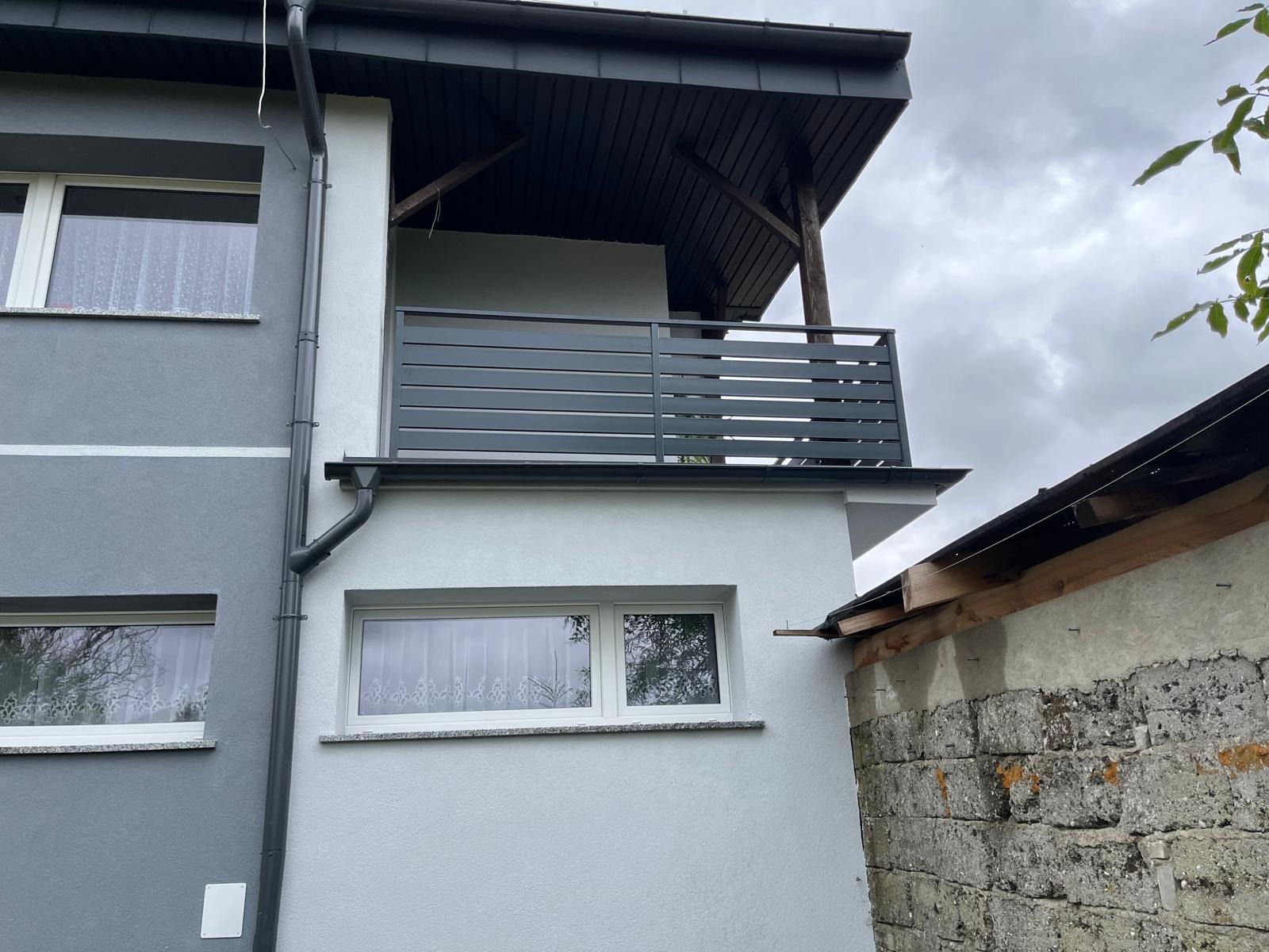 Szara, pozioma balustrada balkonowa z metalowych profili, zamontowana na jasnym budynku z szarą elewacją. Widok z dołu, pochmurne niebo w tle.