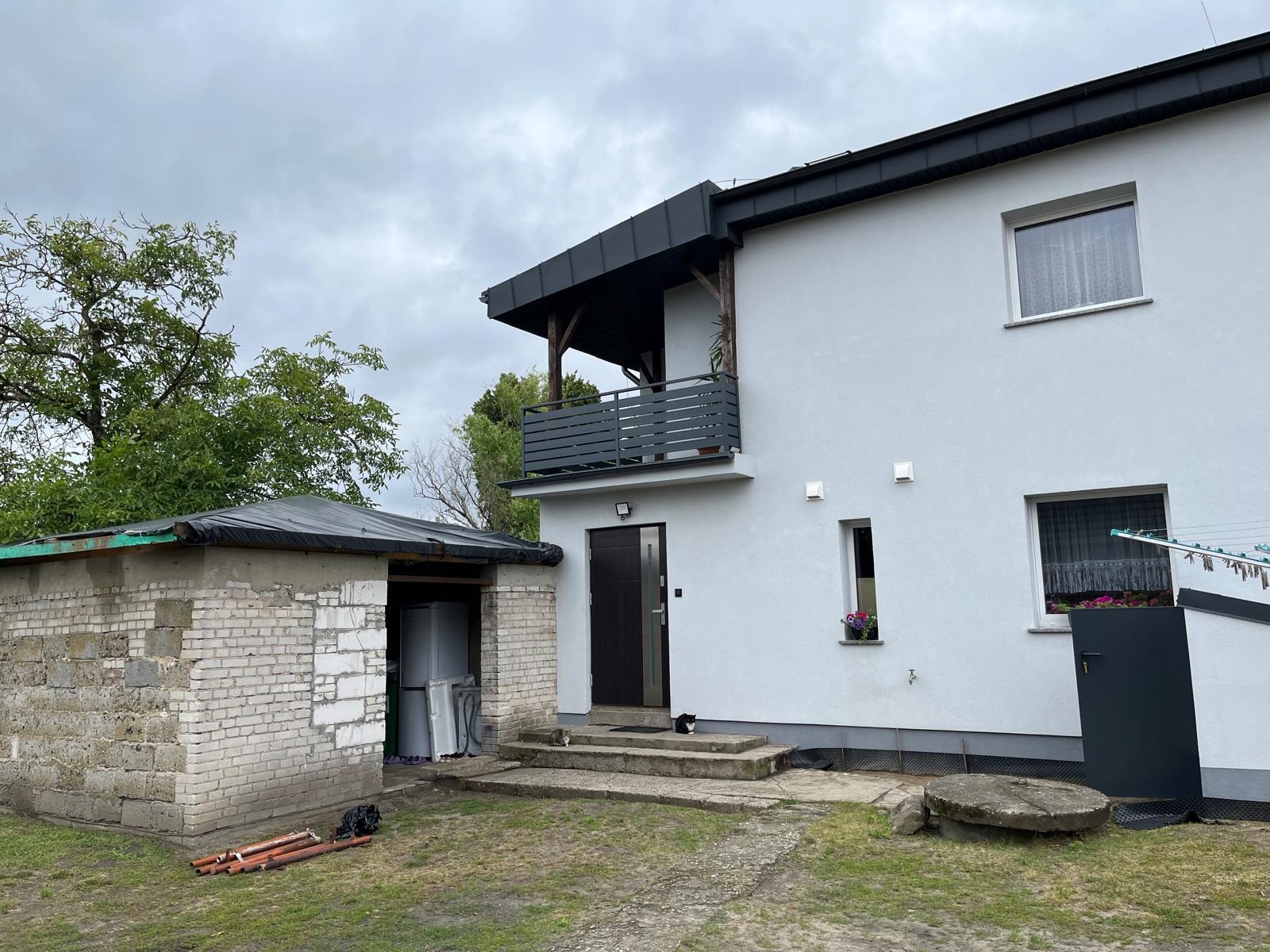 Nowoczesny balkon z szarą, poziomą balustradą, drewnianymi podporami i czarnym zadaszeniem, przylegający do białego domu z szarą stolarką okienną.