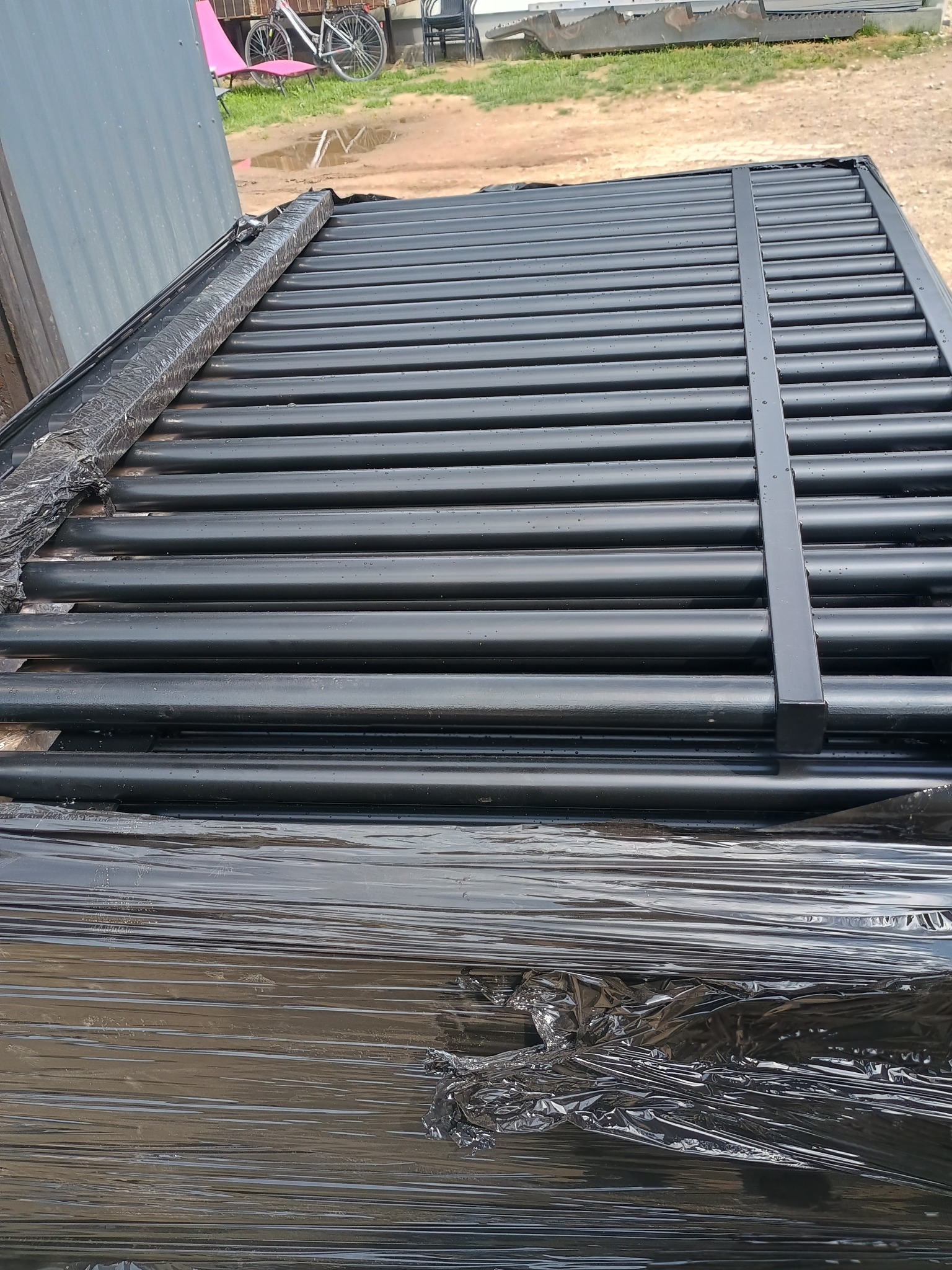 Czarne, metalowe elementy konstrukcyjne, prawdopodobnie fragmenty ogrodzenia, zabezpieczone folią stretch, czekające na montaż. Widoczne rzędy rurek i poprzeczki.