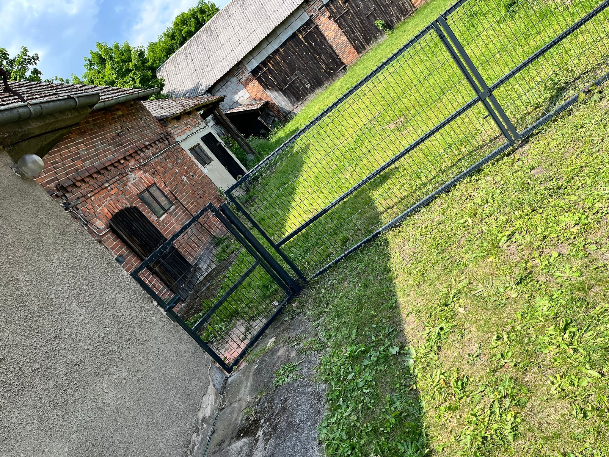 Nowoczesne, metalowe ogrodzenie panelowe w kolorze grafitowym, oddzielające posesję z budynkami gospodarczymi od trawnika. Widok z boku, w tle ceglany budynek.