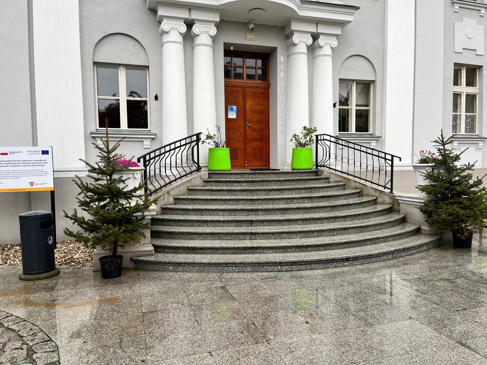 Granitowe schody z czarną, metalową balustradą prowadzą do wejścia budynku z kolumnami. Po bokach schodów stoją donice z roślinami.