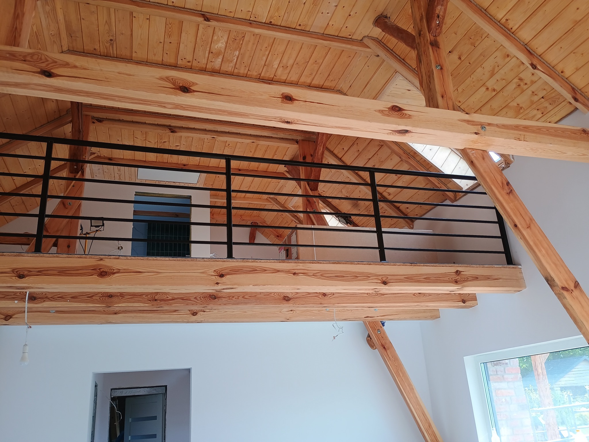 Wnętrze z drewnianą konstrukcją stropu i czarną, metalową balustradą na antresoli. Widoczne elementy drewniane i fragment okna. Ujęcie z dołu.