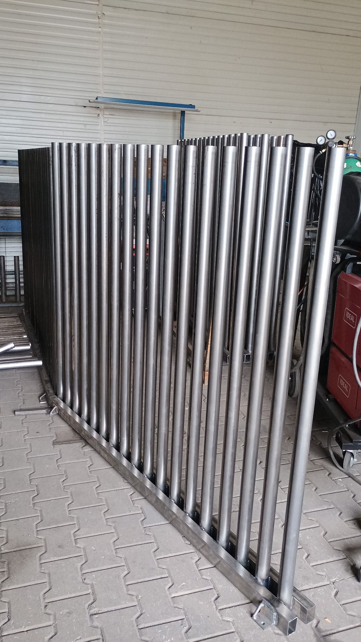 Surowa, metalowa konstrukcja z rur i profili, prawdopodobnie fragment balustrady lub ogrodzenia, w warsztacie. Widoczne ślady spawania i obróbki.
