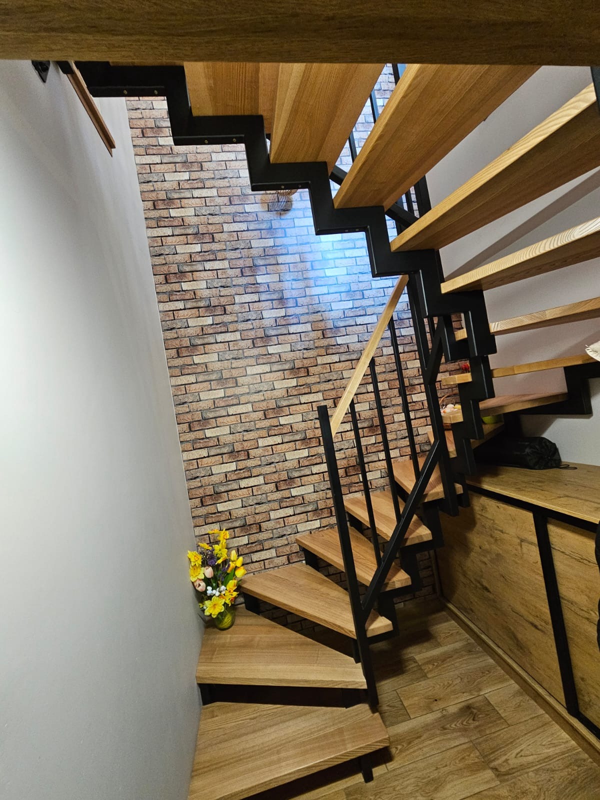 Nowoczesne schody drewniane z czarną metalową konstrukcją, widok z dołu, z kwiatami na pierwszym stopniu i ścianą z cegły w tle. Minimalistyczny design.