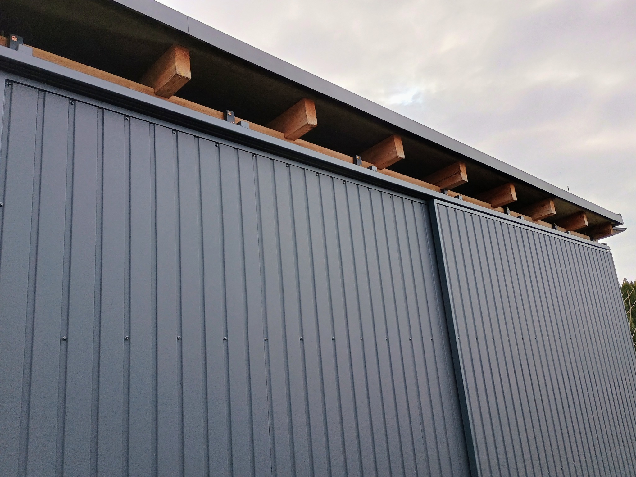Szara, metalowa konstrukcja z widocznymi drewnianymi elementami pod dachem, ukazana pod kątem z dołu. Widoczne łączenia blachy i fragment nieba.