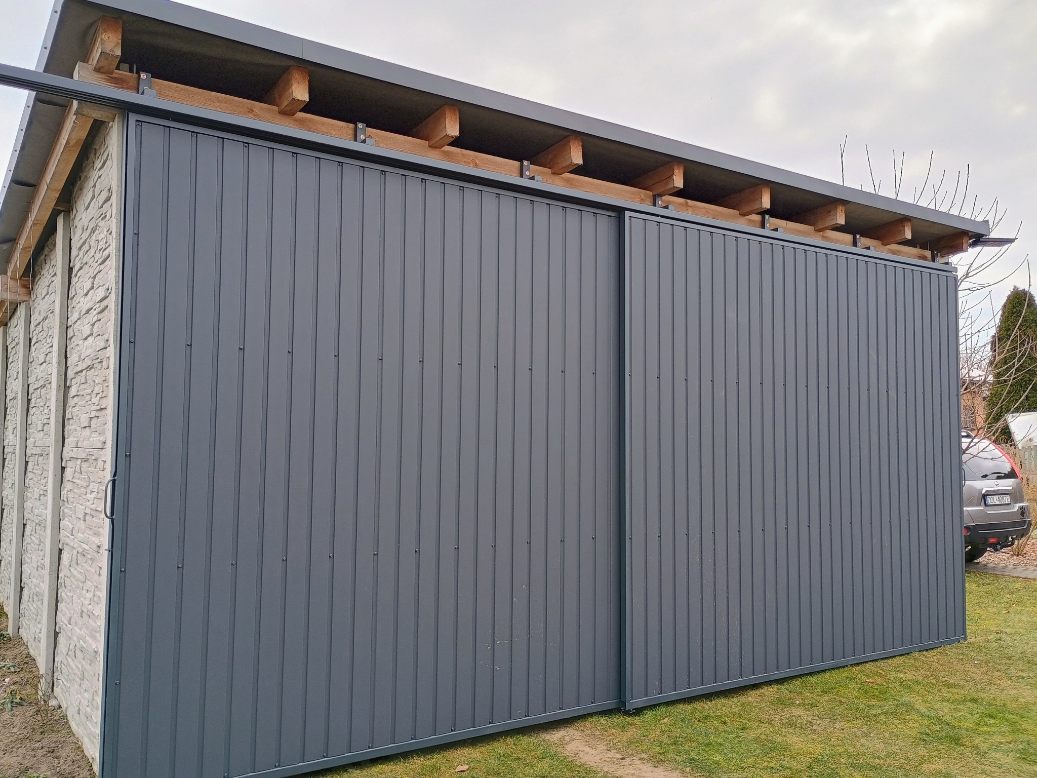 Szara, metalowa brama przesuwna z pionowymi panelami, zamontowana na betonowym murku. Widoczne drewniane elementy konstrukcyjne dachu i fragment samochodu w tle.