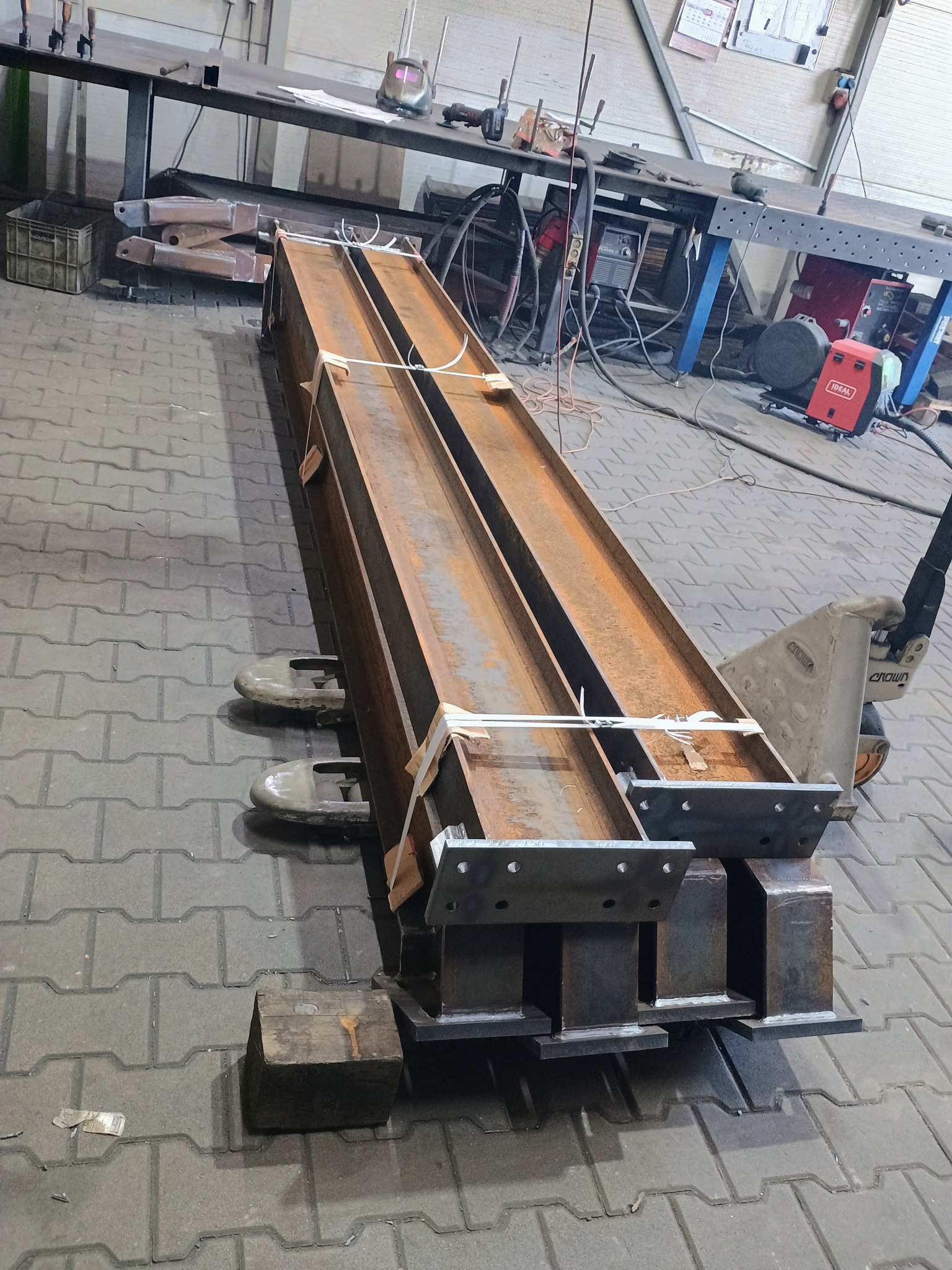 Dwie stalowe belki dwuteowe z przyspawanymi elementami, spięte taśmą, leżą na wózku paletowym w warsztacie. Widoczne spawarki i narzędzia na stołach w tle.