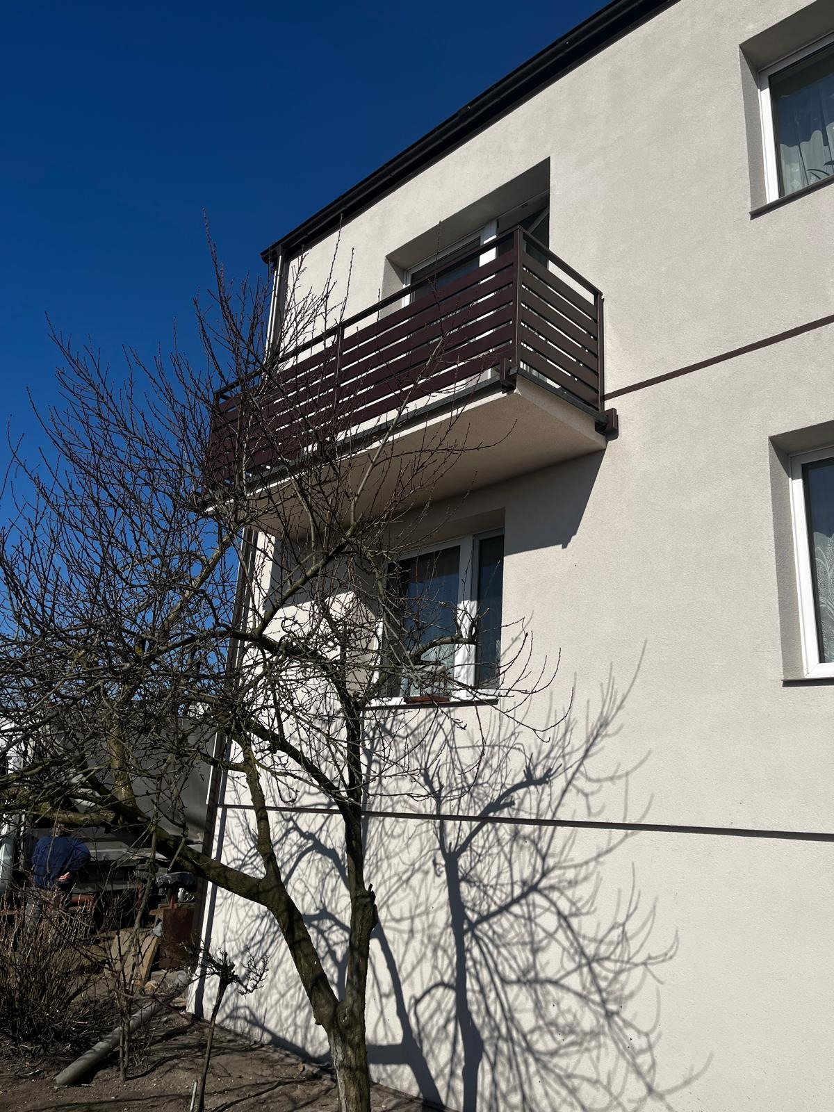 Balkon z brązową, nowoczesną balustradą na tle jasnej elewacji budynku i błękitnego nieba, z drzewem bez liści w pierwszym planie, rzucającym cień na ścianę.