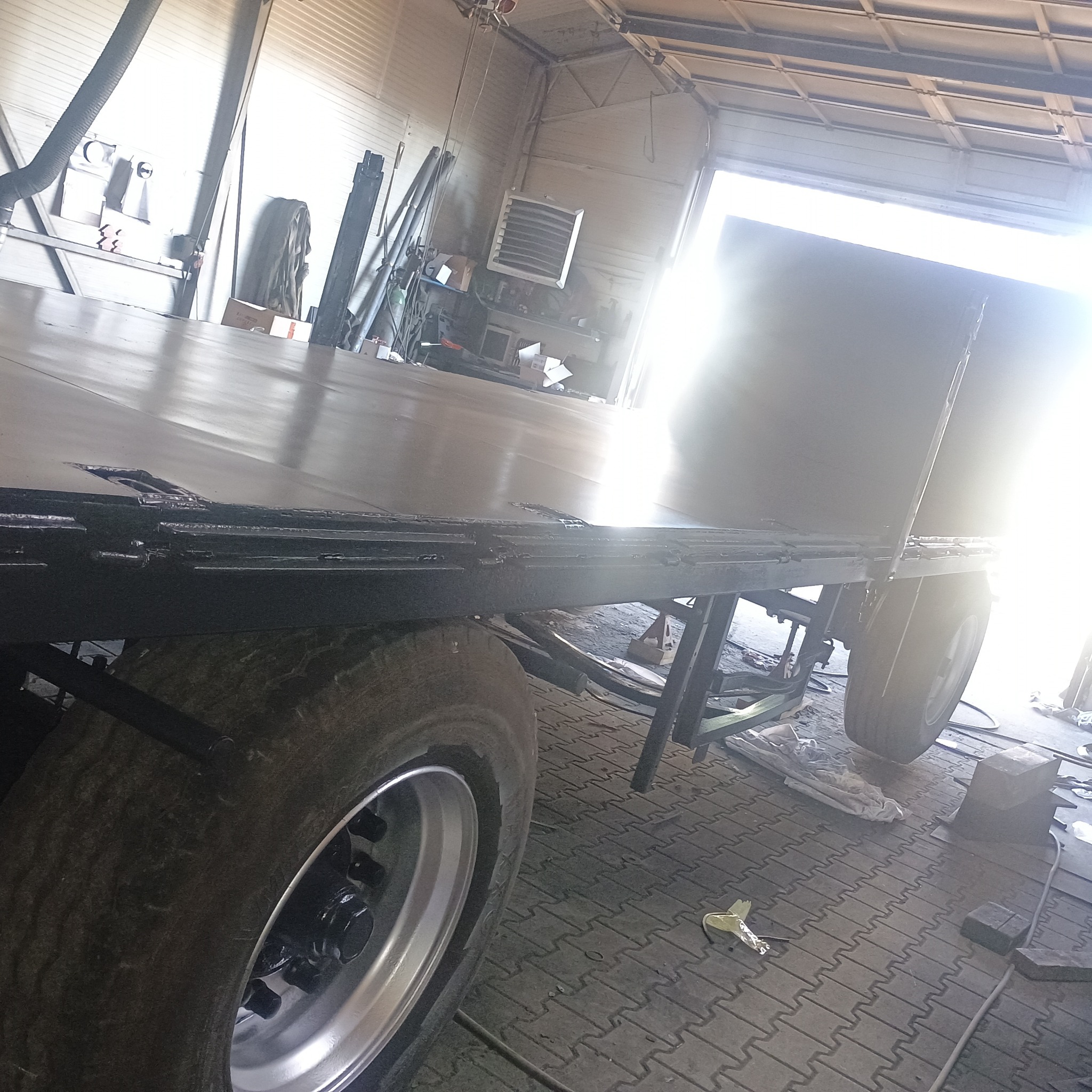 Metalowa konstrukcja przyczepy w warsztacie, widoczne spawane elementy i koło. Jasne światło w tle. Podłoga wyłożona kostką brukową.