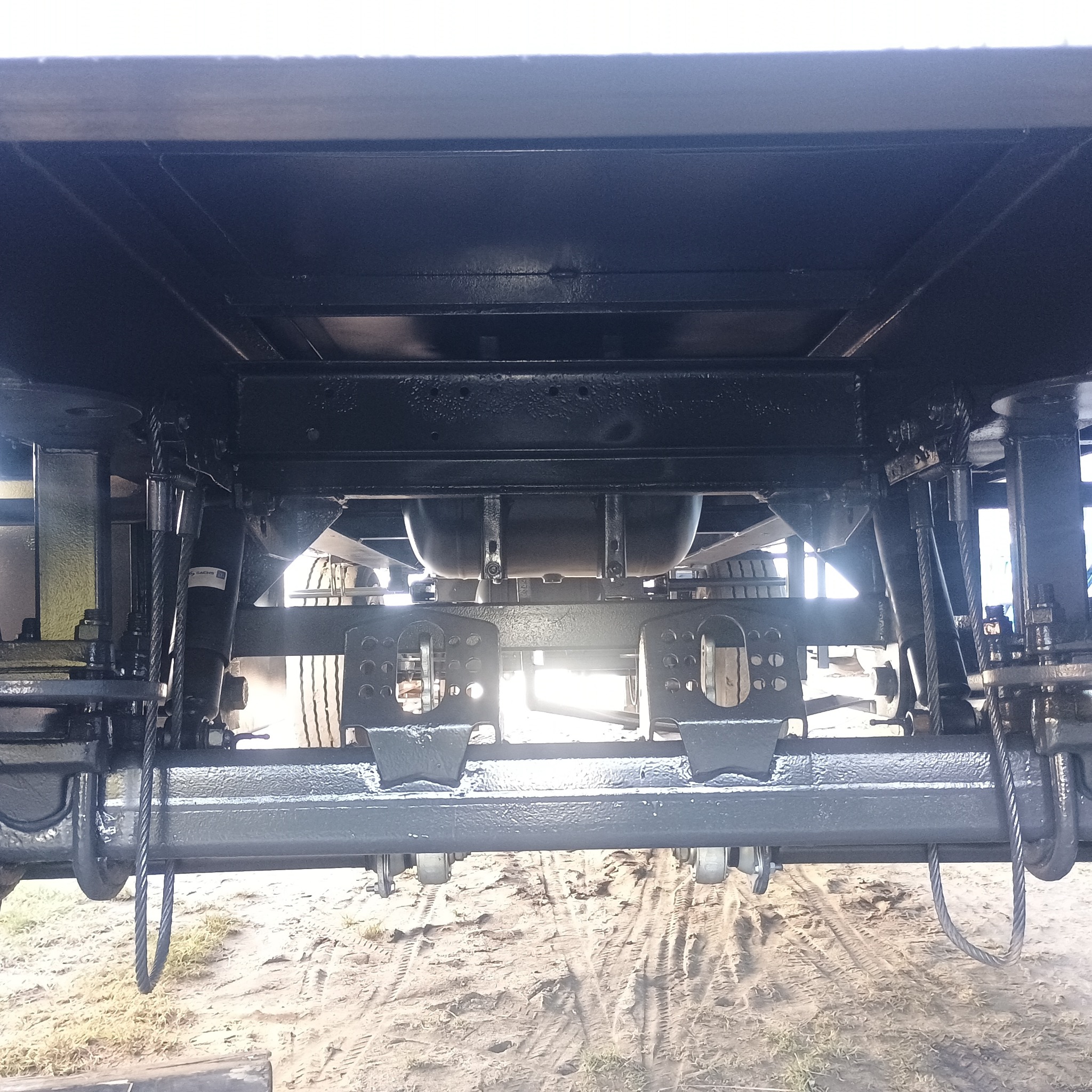 Spawana konstrukcja podwozia przyczepy z widocznymi elementami zawieszenia i przewodami, pomalowana na ciemny kolor, sfotografowana z dołu.