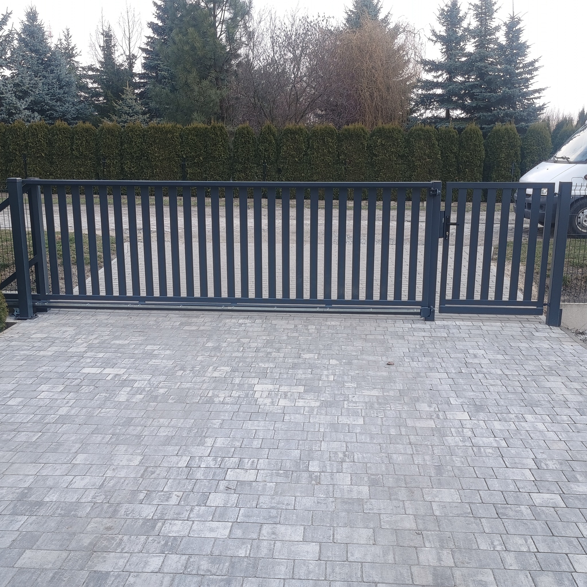 Nowoczesna, przesuwna brama wjazdowa z pionowymi szczeblami, szara kolorystyka, widoczny mechanizm otwierania, kostka brukowa na podjeździe, tło z zielonych krzewów i drzew.