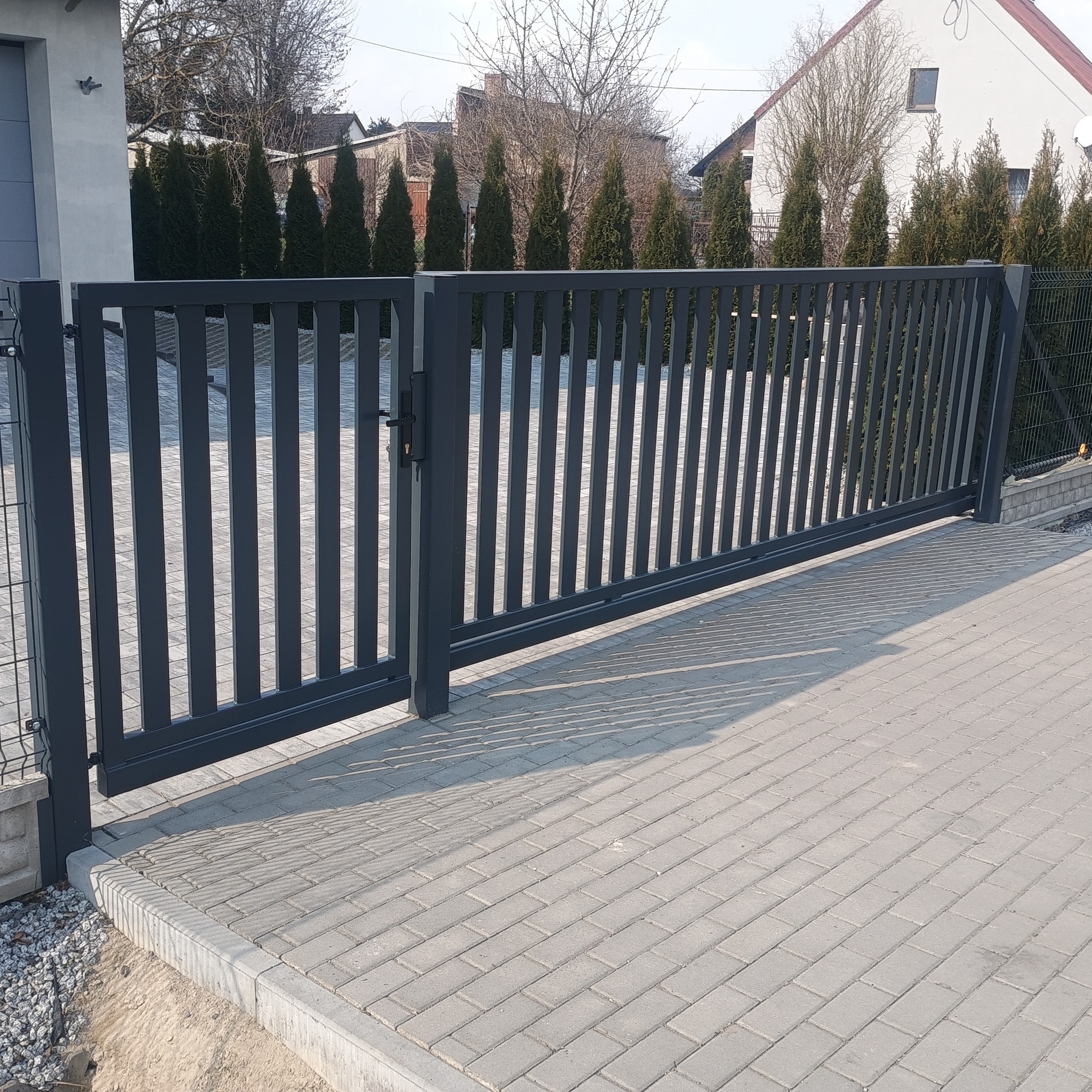 Nowoczesna, przesuwna brama wjazdowa w kolorze grafitowym, z pionowymi szczeblami, zamontowana na posesji z kostką brukową i zielonymi tujami w tle.
