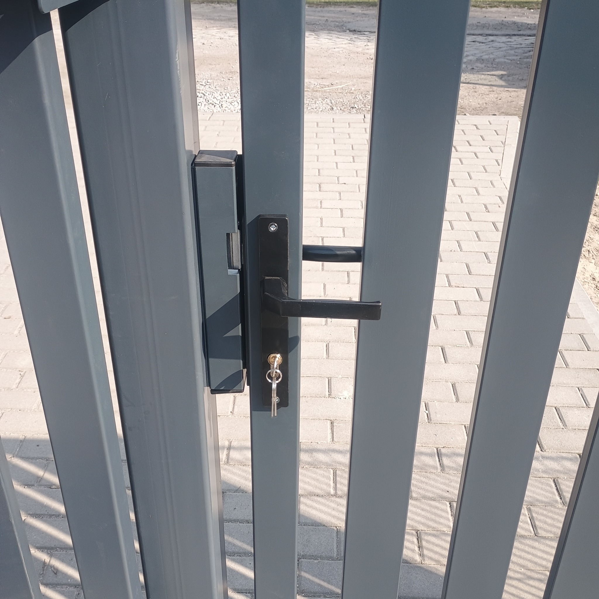 Szara furtka z czarną klamką i kluczem w zamku, widok z bliska na detal. W tle kostka brukowa i żwir.