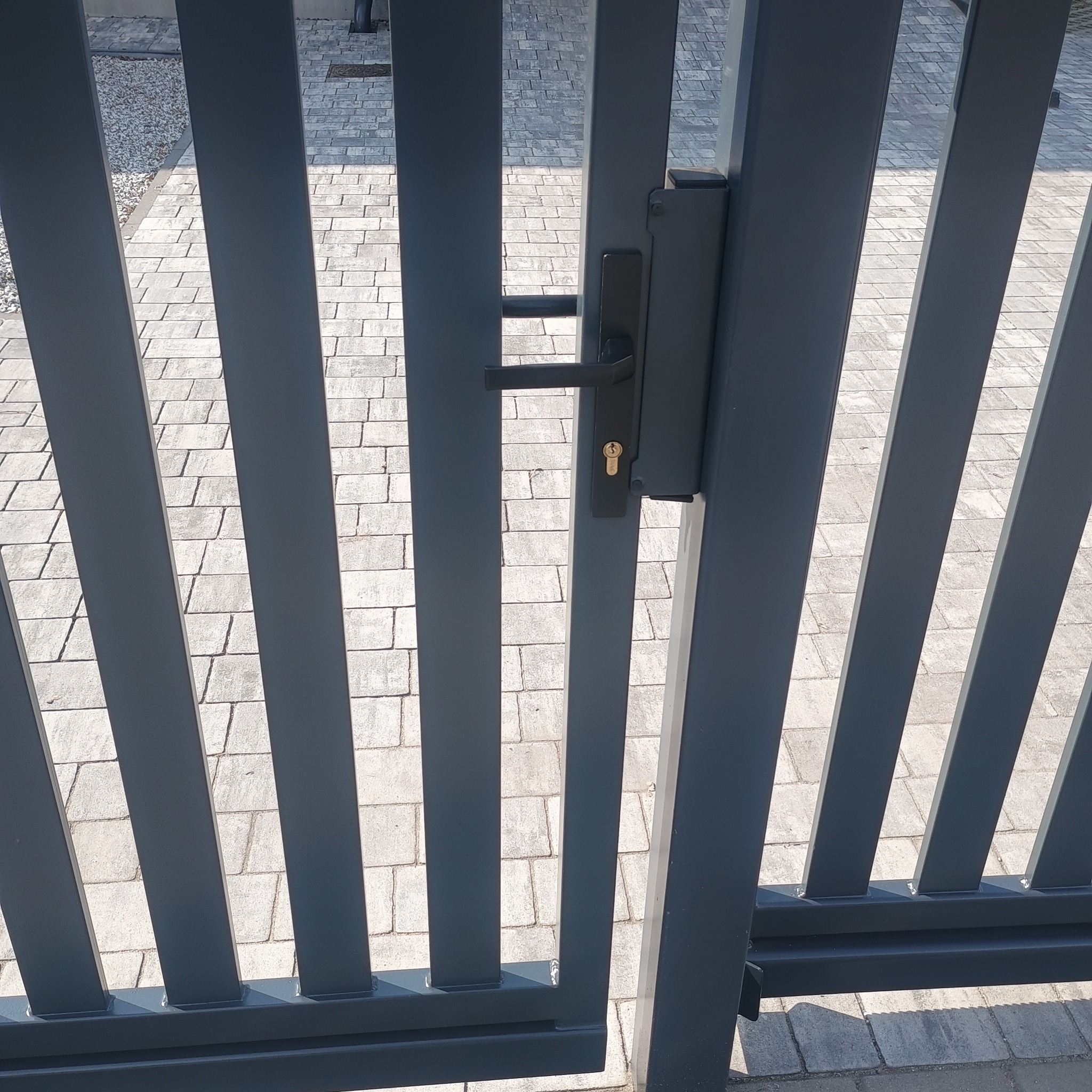 Nowoczesna, grafitowa brama wjazdowa z pionowymi profilami, widok z bliska na zamek i klamkę. Tło stanowi szara kostka brukowa.