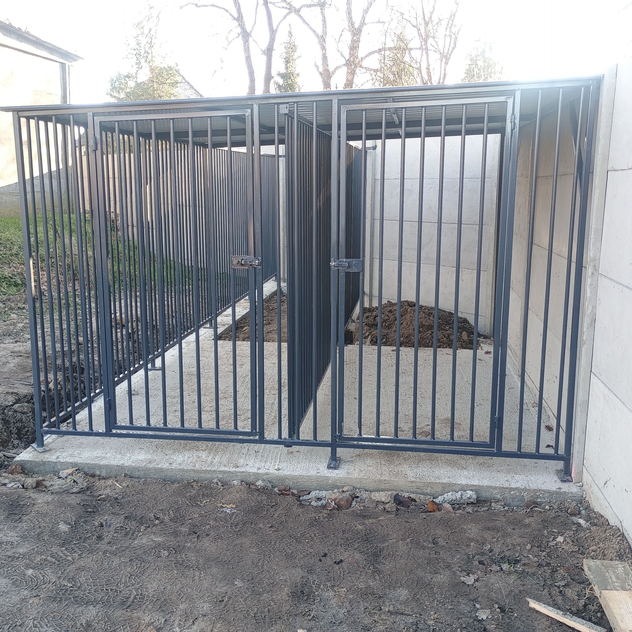 Metalowa konstrukcja z prętów, prawdopodobnie kojec, z dwiema zamykanymi przestrzeniami, na betonowej podstawie, z ziemią w środku. Widoczne ślady budowy.